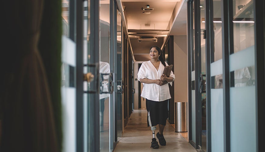 A woman of Indian descent walking through an office holding a tablet. She has a prosthetic leg.