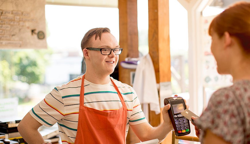 A young person with a disability wears an apron and serves a customer at a 