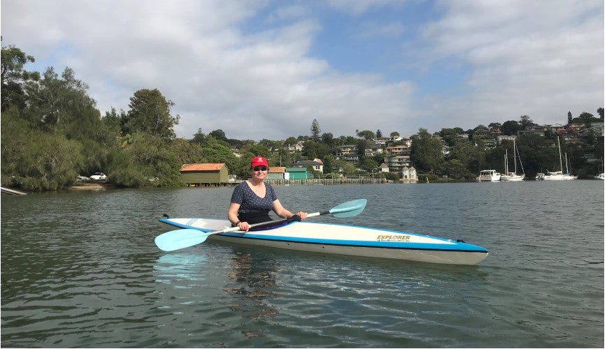 Image of Jane in a white Kayak with blue trimming. Jane is wearing a red cap and sunglasses. She is on the water with trees in the background and the sky is blue with clouds.