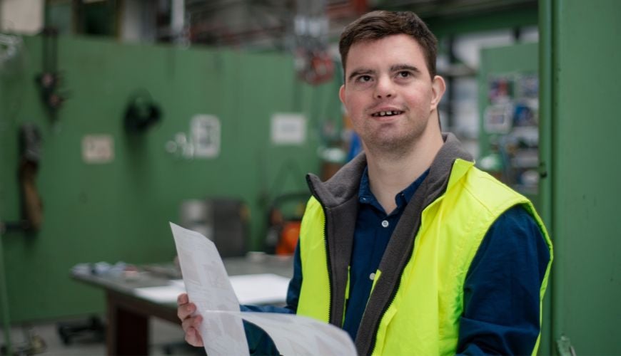 A man with Down Syndrome in an industrial working environment holding some paperwork and wearing a safety vest.