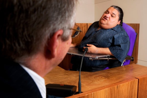 A person in a wheelchair sits giving testimony from the witness box of a courtroom