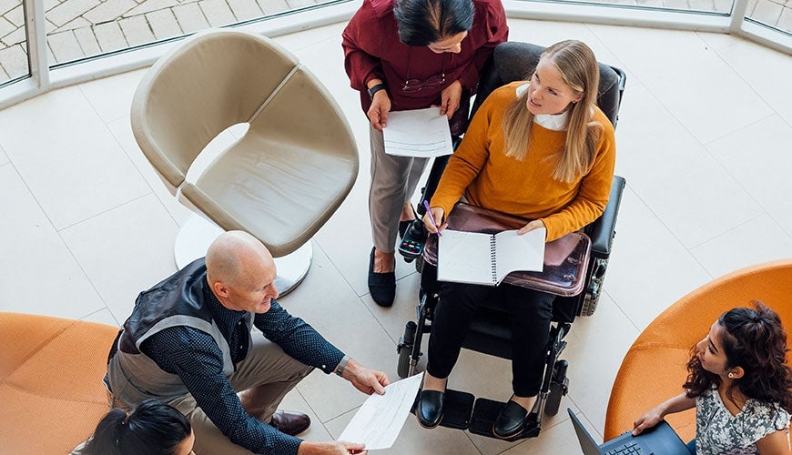 A bird’s eye view of a group of people sitting in a circle with laptops and notebooks. Some people are on couches, and others are on chairs. One woman is in a wheelchair.