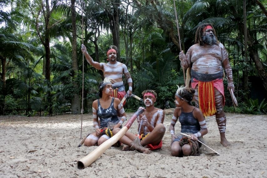 First Nations children in traditional dress, surrounded by elders, sitting on the sand holding traditional instruments like didgeridoo