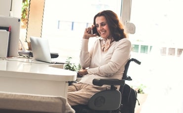 A person in a wheelchair sits at their desk while talking on the phone smiling