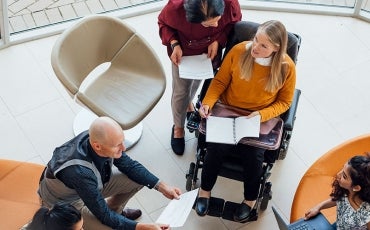 people sitting in a circle with laptops and notebooks, one of whom is in a wheelchair