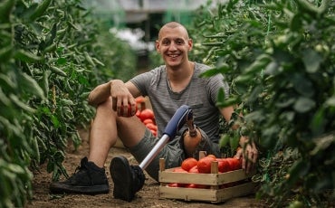 A person with a prosthetic leg sits between rows of crops, smiling and picking tomatoes