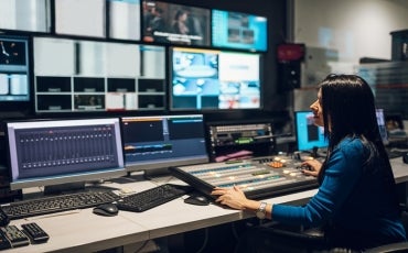 Middle aged woman in blue shirt sitting at a desk using equipment in control room on a TV station