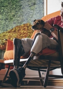 A young man is sitting in an armchair patting a dog. He is wearing prosthetic lower legs.