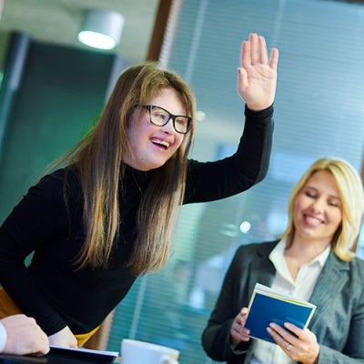 Two colleagues are raising hands for a high five. One is partially out of frame and the other is a young woman with long blonde hair and Down Syndrome.  A male colleague is smiling and  a woman is also smiling and looking down at a book.