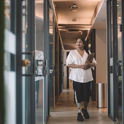 A woman of Indian descent walking through an office holding a tablet. She has a prosthetic leg.