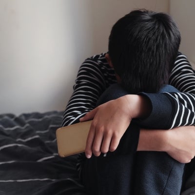 Asian teenager boy hugging his knee in his bedroom with smartphone.